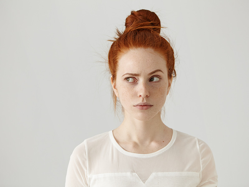 Hmm. Let me think. Studio shot of cute redhead girl with hair knot and freckles looking sideways with thoughtful and sly expression, raising one brow as if having good idea, planning something